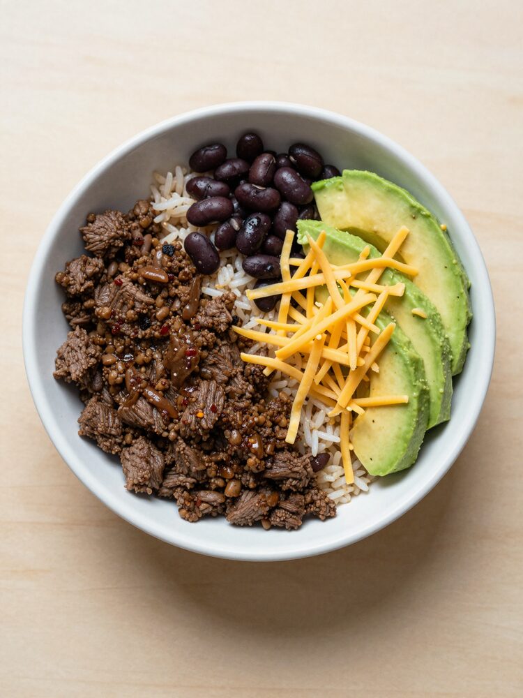 Meal prep bowl with seasoned ground beef, rice, black beans, sliced avocado and shredded cheese
